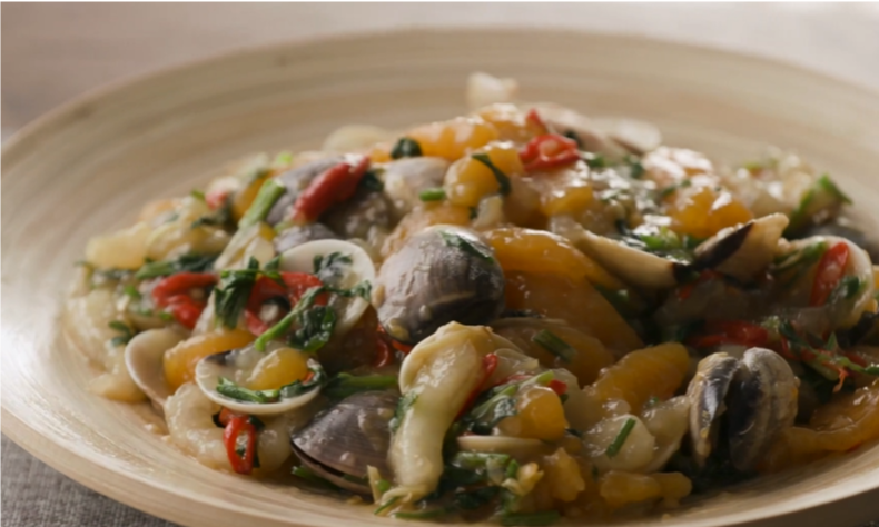 A wooden bowl contains a colorful stir-fry with clams, sliced vegetables, leafy greens, and red chili peppers.